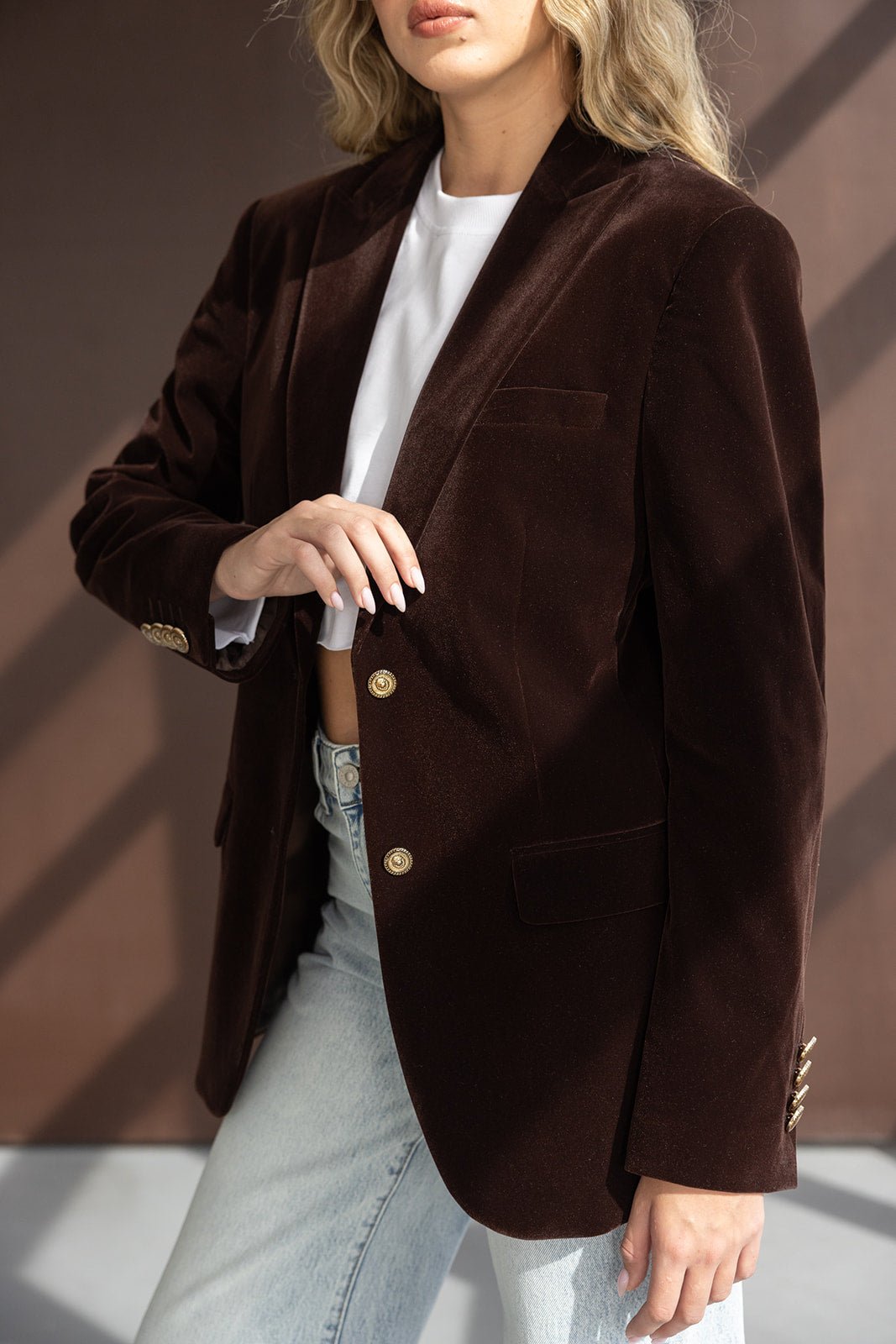 אישה עם בלייזר קטיפה חום כהה וכפתורים זהב, לובשת ג'ינס אופנתי וחולצה לבנה