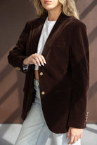 אישה עם בלייזר קטיפה חום כהה וכפתורים זהב, לובשת ג'ינס אופנתי וחולצה לבנה