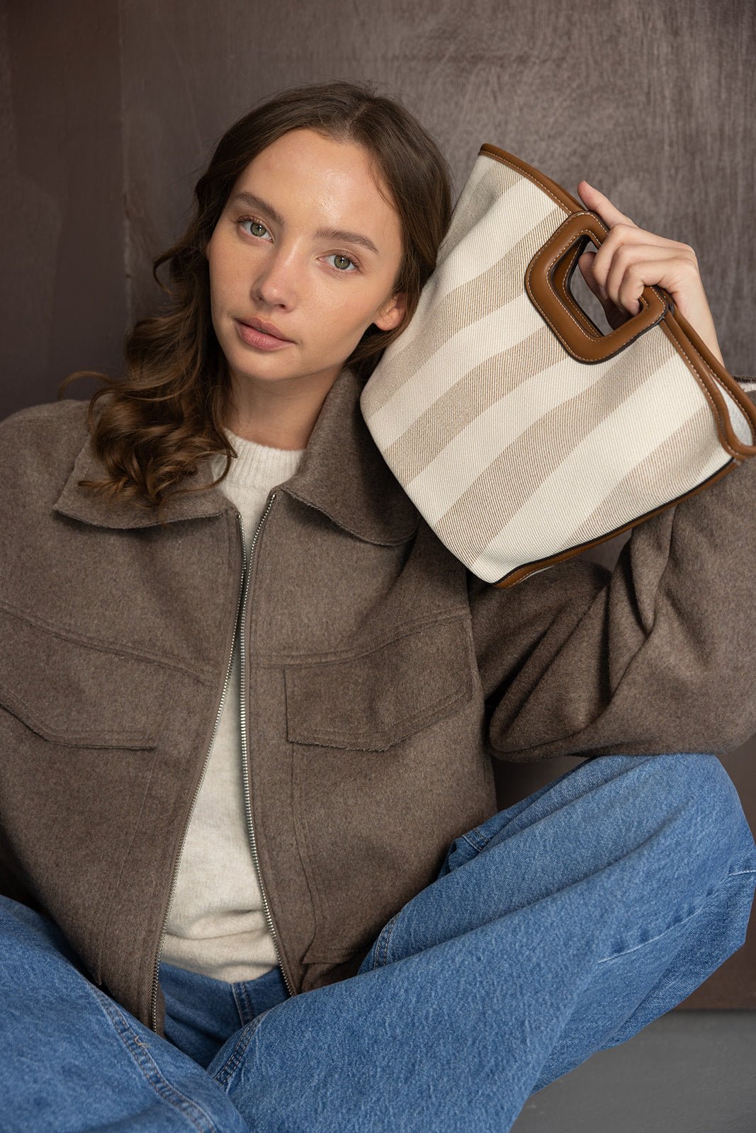 דוגמנית יושבת עם מעיל חום אופנתי, ג'ינס ותיק יד אופנתי בפסים בז' קרם