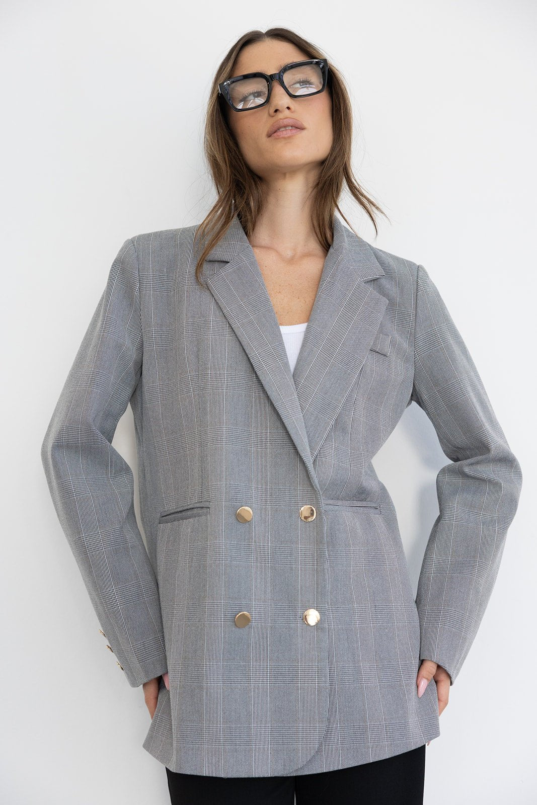 אישה עם בלייזר מחויט משובץ אפור, כפתורים זהב, משקפי אופנה גדולים, רקע לבן