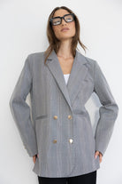 אישה עם בלייזר מחויט משובץ אפור, כפתורים זהב, משקפי אופנה גדולים, רקע לבן