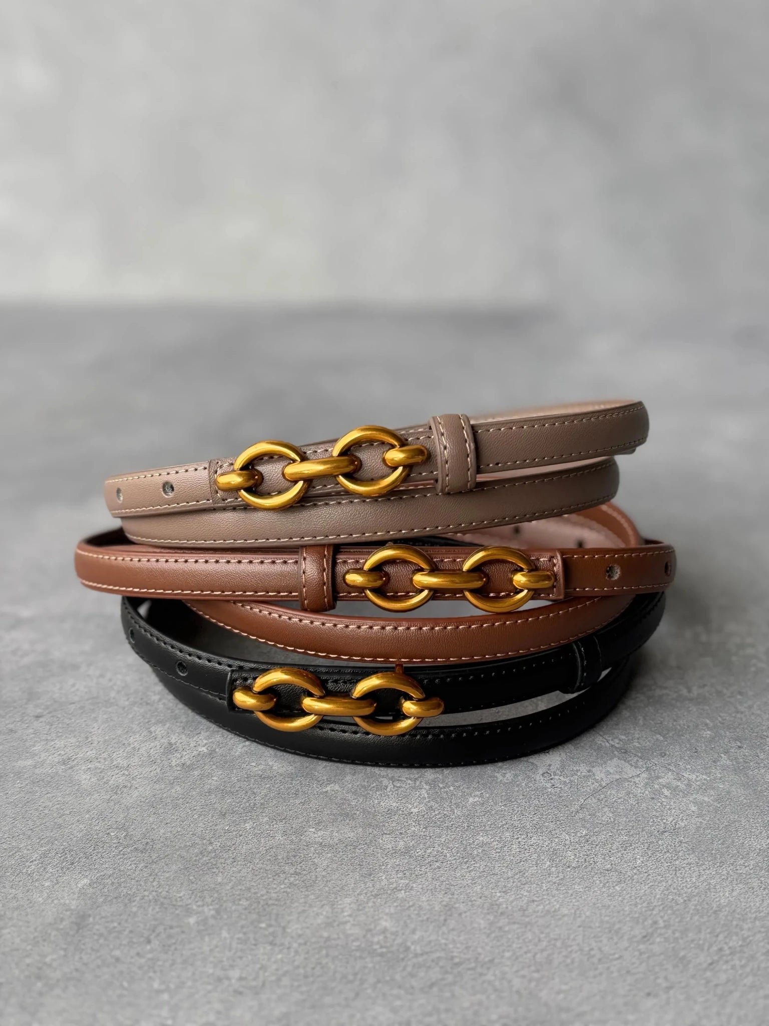 שלושה חגורות נשים בצבע חום, בז' ושחור עם אבזם זהב, צילום על רקע אפור