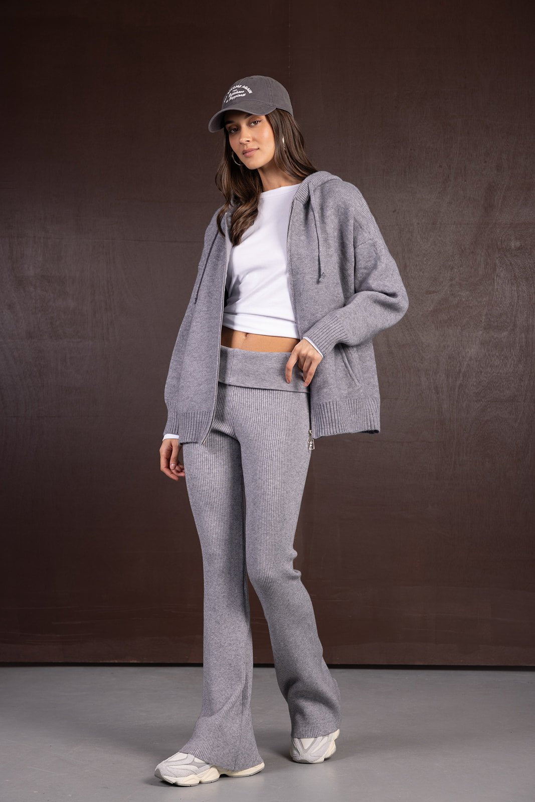 דוגמנית לובשת סט טרנינג אפור עם קפוצ'ון, מכנסיים סרוגים, חולצה לבנה וכובע מצחייה אופנתי.