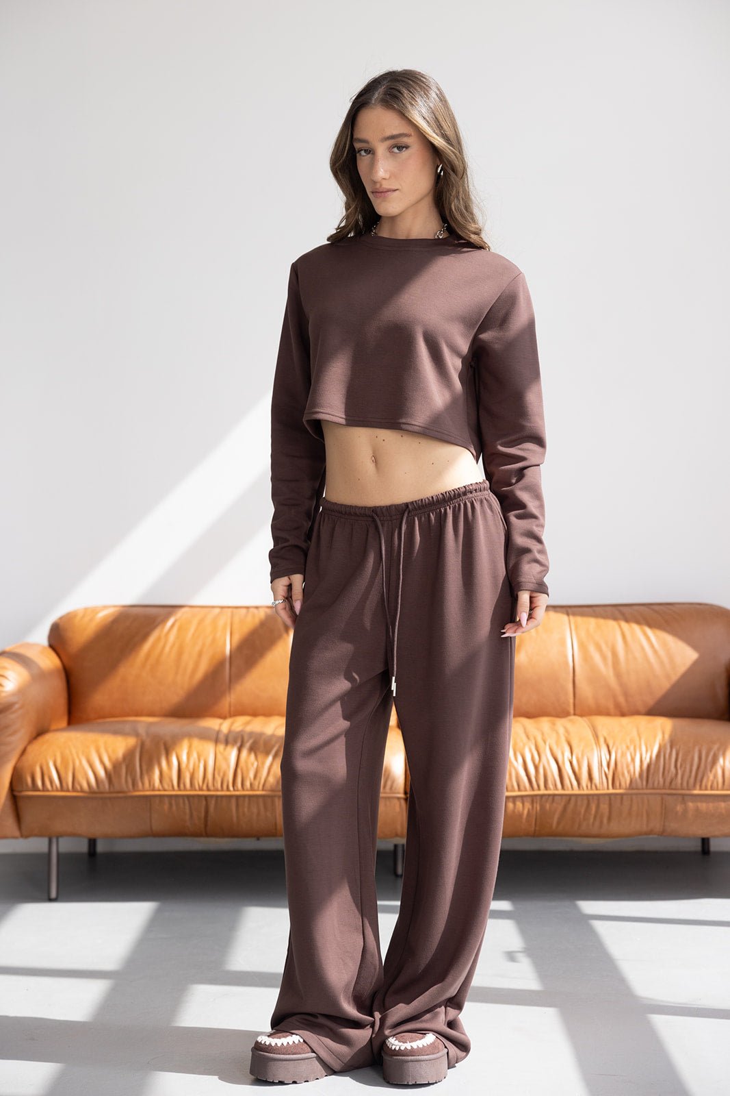 אישה לובשת סט טרנינג חום אופנתי עם חולצה בטן מכנסיים רחבים ונעליים, עומדת בסלון מודרני