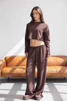 אישה לובשת סט טרנינג חום אופנתי עם חולצה בטן מכנסיים רחבים ונעליים, עומדת בסלון מודרני