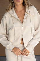 אישה לובשת סט טרנינג אופנתי בצבע בז', קפוצ'ון עם רוכסן וכיתוב, מתאים לאופנת רחוב ונשים צעירות