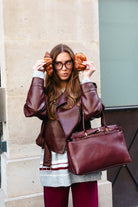 אישה עם משקפיים ולבוש אופנתי בצבע בורדו, מחזיקה בגטים ליד הראש, תיק עור בורדו.