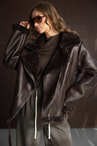 אישה עם מעיל דמוי עור חום, חולצה חומה, משקפי שמש אופנתיות ומכנסיים רחבים