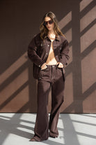 אישה לובשת חליפת ג'ינס חומה רחבה עם תפרים לבנים, משקפי שמש, עומדת ברקע חום מואר