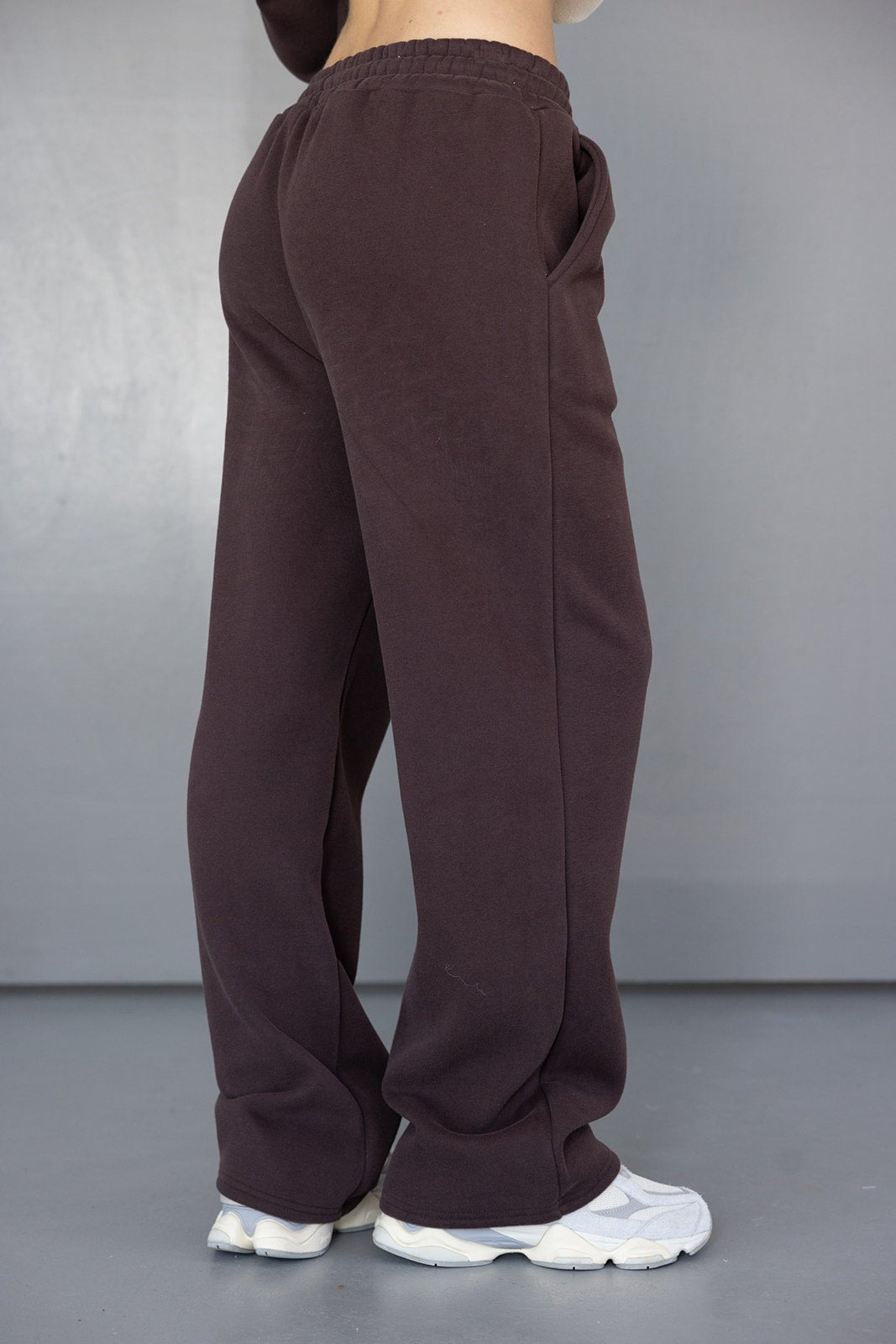 מכנסי טרנינג רחבים לנשים בצבע חום כהה עם גומי במותן, נעלי סניקרס לבנות, רקע אפור