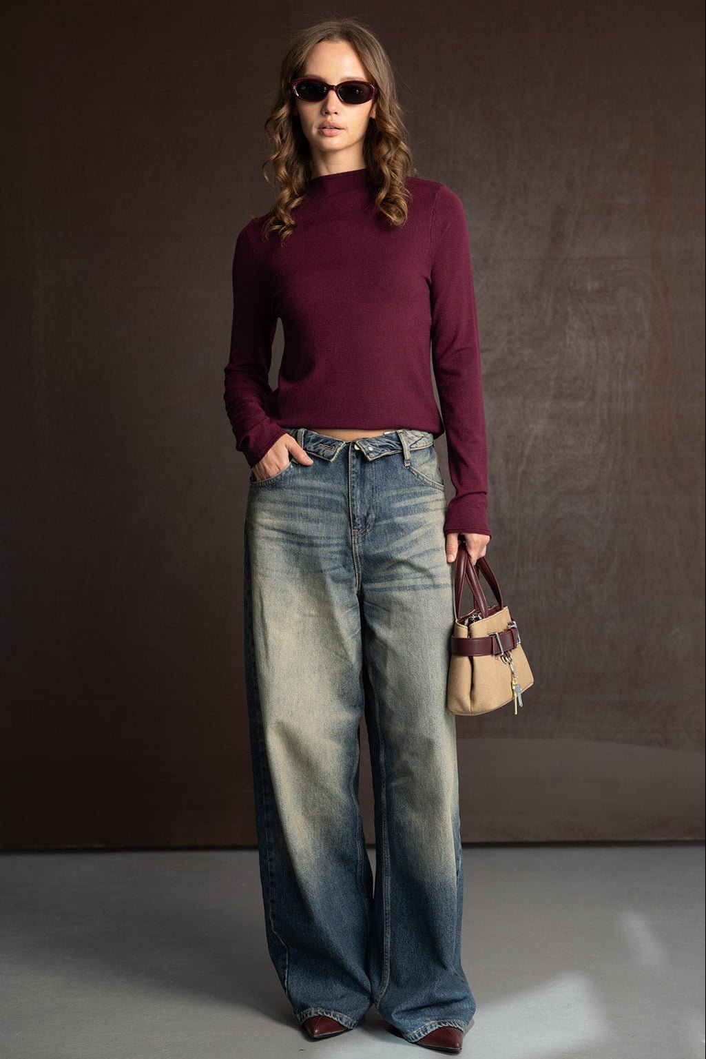 אישה עם חולצה בורדו ארוכה, ג'ינס רחב משופשף, משקפי שמש ותיק יד קטן לאופנת נשים ונערות