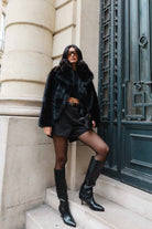 אישה עם מעיל פרווה שחור, חולצה שחורה, מכנסיים קצרים, גרביונים ומגפיים גבוהים אופנתיים