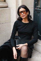 אישה עם משקפי שמש גדולים, לובשת חולצה שחורה וחצאית, מחזיקה תיק קלוע, יושבת ברחוב אופנתי