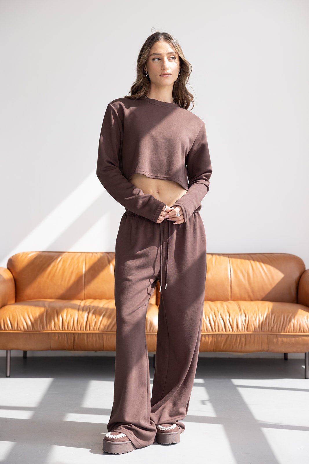 דוגמנית לובשת סט טרנינג חום מחויט לנשים, מכנסיים רחבים וחולצה קצרה, רקע סלון מודרני