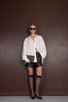 אישה עם חולצה לבנה רחבה, מכנסי עור שחורים, משקפי שמש ושקית קלאץ' אופנתית