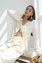 אישה יושבת על ספסל בלבוש אופנתי בגוון שמנת, חולצה עם שרוך ומכנסיים רחבים, רקע מואר