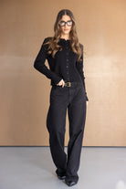 אישה עם שיער גלי ולבוש אופנתי לובשת קרדיגן שחור, מכנסיים שחורים ומשקפיים עבים, עומדת ברקע נייטרלי