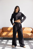 אישה לובשת סט טרנינג שחור לנשים, חולצה קצרה ומכנסיים רחבים, בסלון מודרני מואר