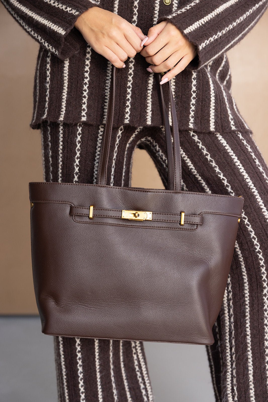 תיק יד חום מעור לנשים עם אבזם זהב, מונף על ידי דוגמנית בלבוש חורפי מפוספס