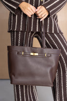 תיק יד חום מעור לנשים עם אבזם זהב, מונף על ידי דוגמנית בלבוש חורפי מפוספס