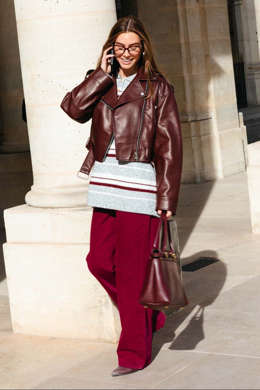 אישה באופנת רחוב עם ג'קט עור בורדו, סוודר מפוספס ומכנסיים רחבים, תיק תואם ומשקפיים