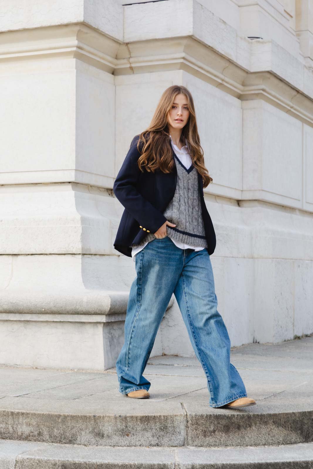 אישה עם בלייזר כחול, סוודר אפור גזור, חולצה לבנה וג'ינס רחב אופנתי עומדת מול מבנה אבן