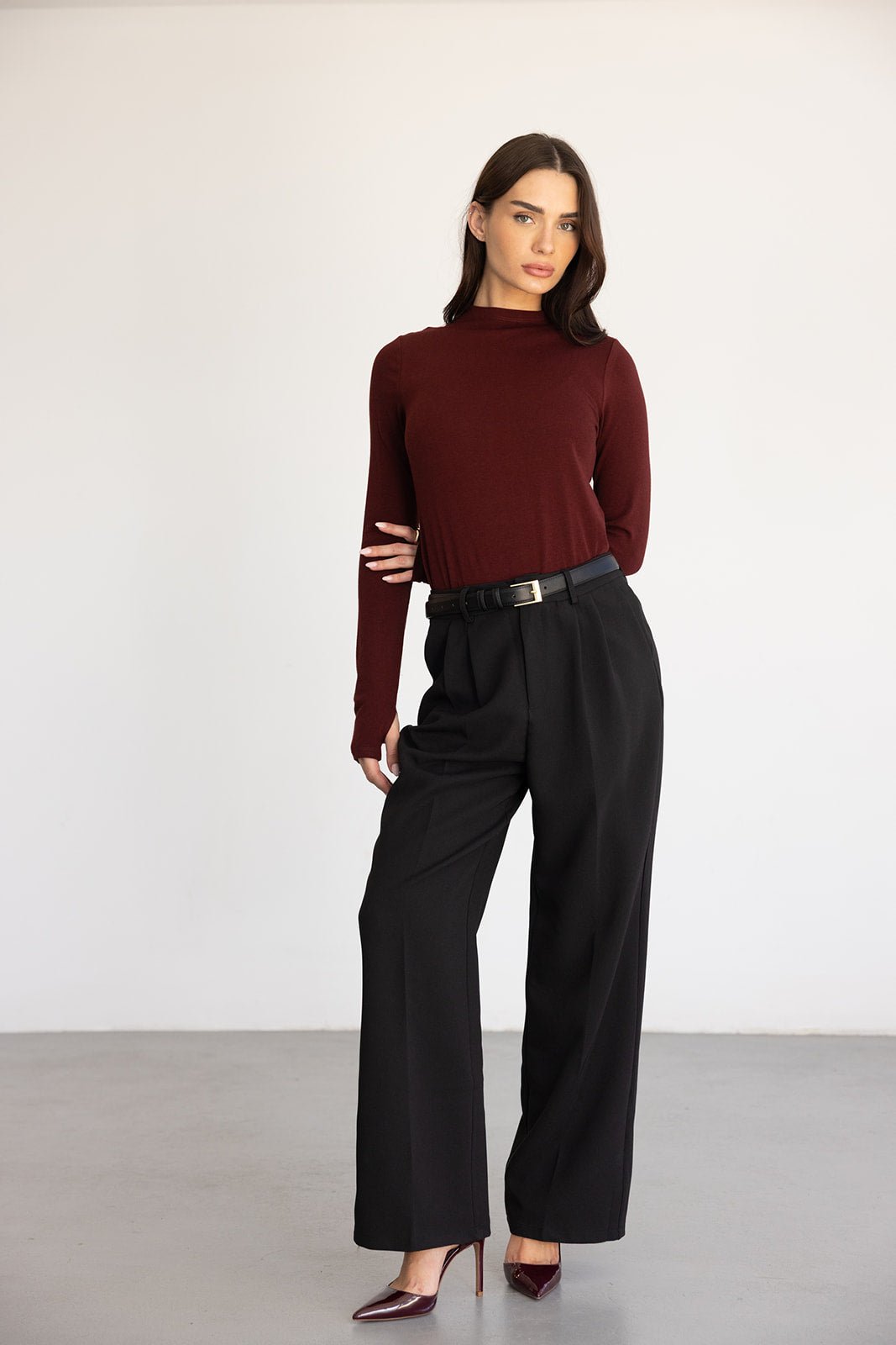 אישה לובשת חולצת בורדו ארוכה, מכנסיים שחורים מחויטים ונעלי עקב, אופנת נשים אלגנטית בעמידה