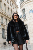 אישה צעירה בלבוש שחור אופנתי, מעיל פרווה, מכנסיים קצרים וחולצה, ברחוב עירוני יוקרתי