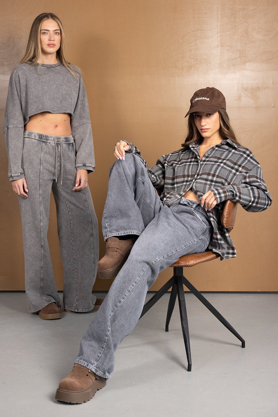 שתי נשים לובשות בגדי אופנה טרנדיים: אחת בחליפת טרנינג אפורה, השנייה בג׳ינס רחב וחולצת פלנל משובצת, נעלי פלטפורמה ומגבעת חומה, צילום על רקע חום