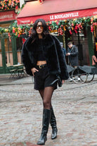 אישה אופנתית בלבוש חורפי שחור, מעיל פרווה, חצאית קצרה ומגפיים גבוהים, ברחוב עירוני