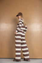 אישה לובשת חליפת פסים חומה ולבנה וכובע חום, עומדת על רקע חום, אופנת נשים צעירה