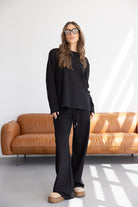 אישה מדגמנת סט טרנינג שחור אופנתי, משקפי שמש, ספה חומה ברקע, אופנת נשים טרנדית