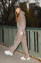 אישה צעירה לבושה בסט טרנינג חום וכובע תואם, צועדת על גשר עירוני עם נעלי ספורט לבנות, אופנת רחוב לנשים