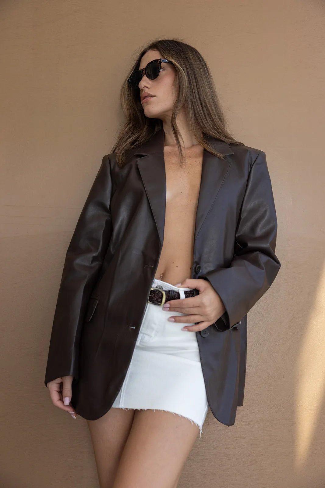 אישה עם ז'קט עור חום, חצאית מיני לבנה ומשקפי שמש אופנתיים, על רקע חום