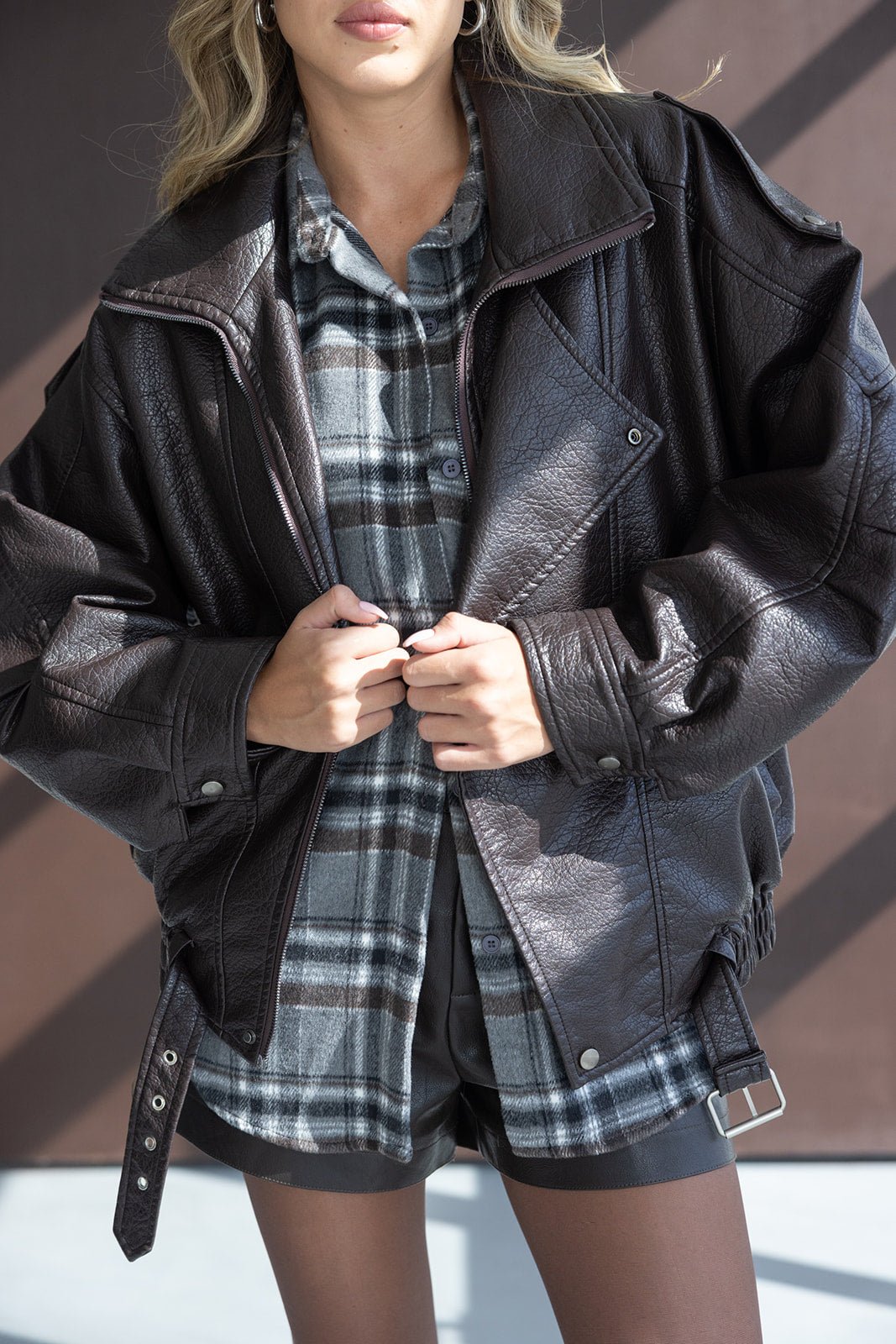 אישה לובשת ג'קט עור חום אוברסייז מעל חולצה משובצת ומכנס קצר אופנתי לנשים