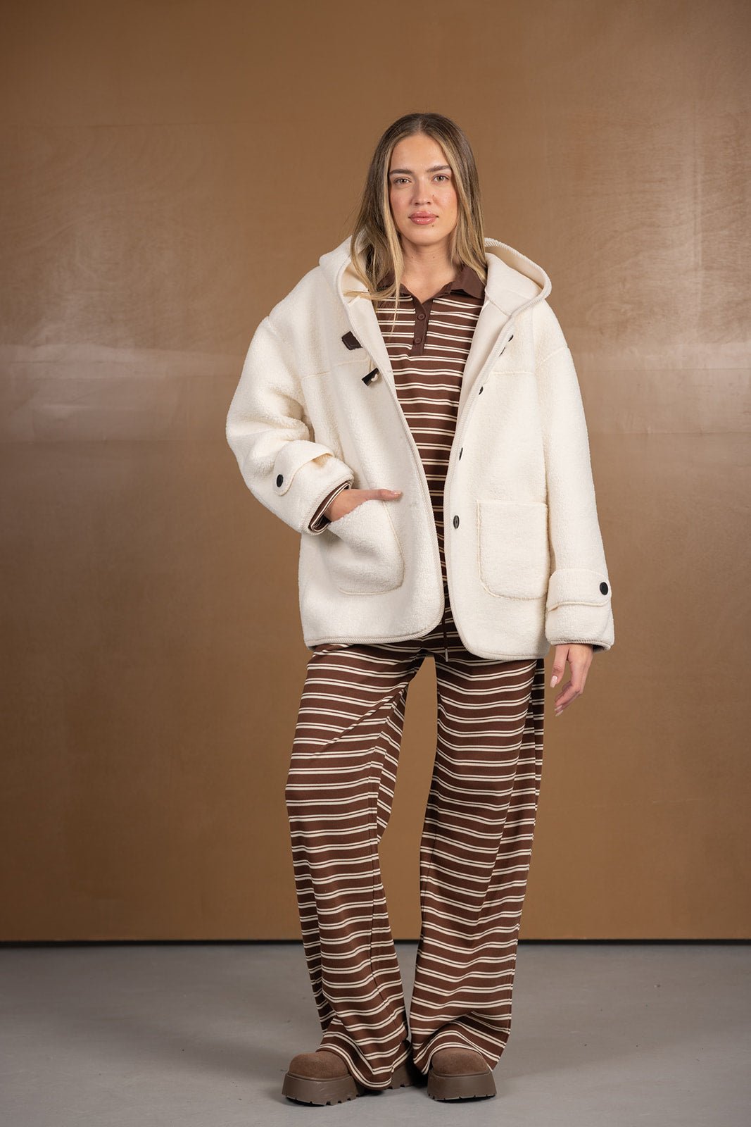 אישה לובשת ג'קט פליז לבן עם קפוצ'ון מעל סט פסים חום ולבן, אופנת חורף לנשים