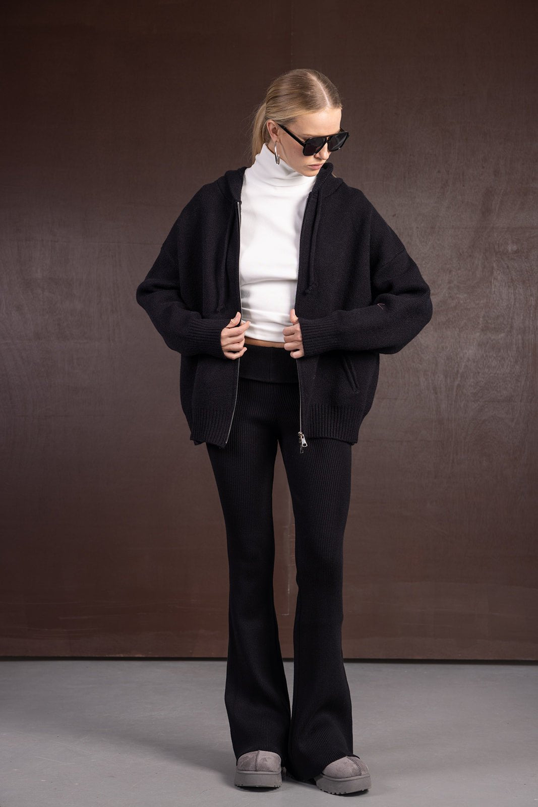 אישה לובשת סוודר רחב שחור, מכנסיים מחמיאים שחורים, טופ גולף לבן ומשקפי שמש אופנתיים