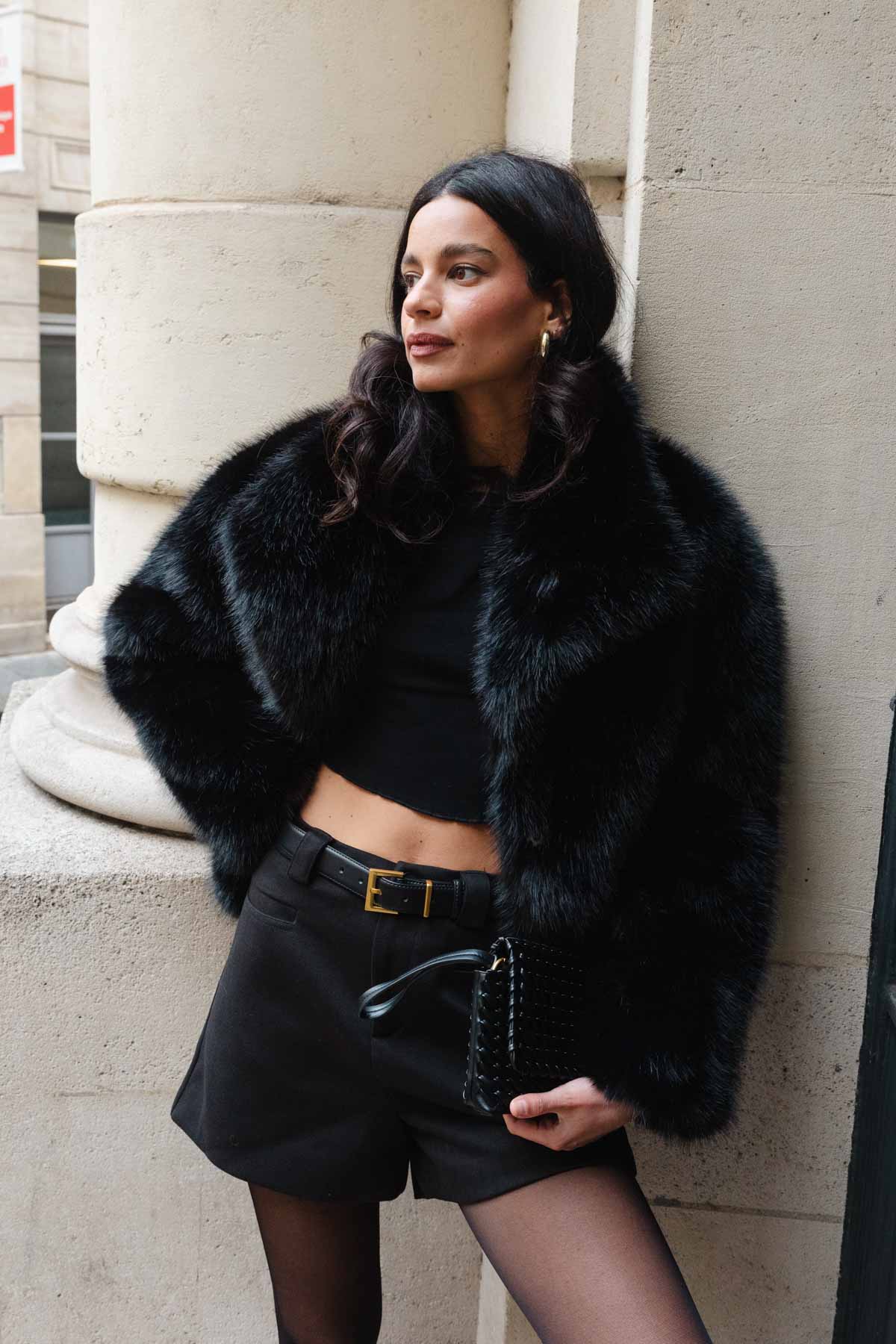 אופנת נשים אלגנטית: אישה עם מעיל פרווה שחור, טופ קצר, מכנסיים קצרים וחגורה, שרשרת זהב ותיק יד שחור