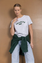 אישה לובשת חולצת טי אפורה עם כיתוב COLLECTS FREEDOM, ג'ינס רחב, וסוודר ירוק קשור במותן, על רקע חום