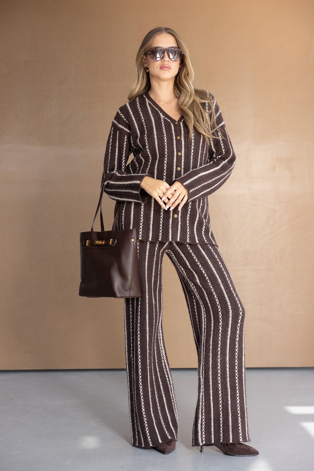 אישה לובשת חליפת נשים חומה מפוספסת, מחזיקה תיק חום ונועלת נעלי עקב, אופנת חורף