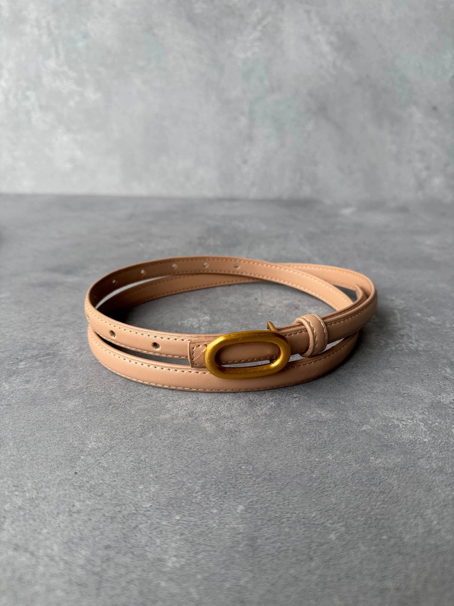 חגורת נשים דקה בצבע בז' עם אבזם מוזהב, מונחת על משטח אפור