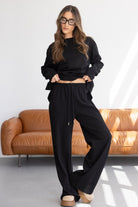 אישה עם שיער ארוך לובשת סט טרנינג שחור עם מכנסיים רחבים בחדר מודרני, אופנת נשים צעירה