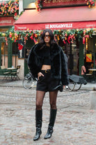 אישה באאוטפיט שחור אופנתי עם מעיל פרווה, מכנסיים קצרים, מגפיים גבוהים ומשקפי שמש בלוק עירוני