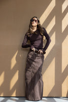 אישה צעירה לובשת חולצה חומה ארוכה וחצאית מקסי חומה עם כיסי צד, משקפי שמש ואבזם חגורה, אופנת נשים טרנדית