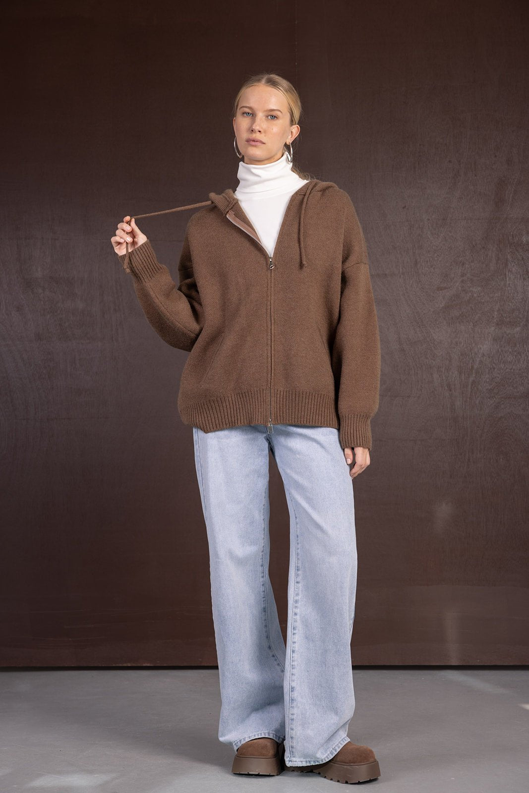 אישה מדגמנת סווטשרט חום עם רוכסן, גולף לבן, ג'ינס רחב ונעלי פלטפורמה חומות, רקע חום