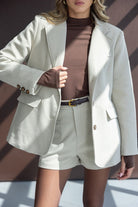 אישה לובשת בלייזר בהיר ומכנסיים קצרים תואמים, עם חולצת גולף חומה וחגורה שחורה אופנתית