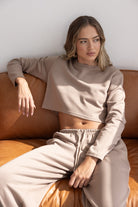 דוגמנית לובשת סט טרנינג בז' אופנתי עם קרופ טופ ארוך שרוול ומכנסי גומי, ישובה על ספה חומה