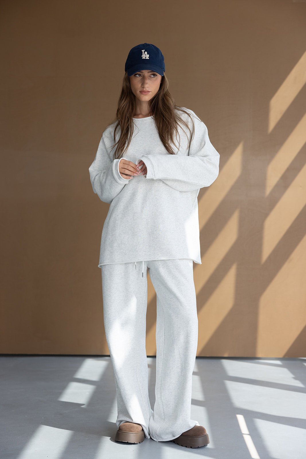 דוגמנית לובשת חליפת טרנינג אפורה לנשים עם כובע כחול ונעלי בית חומות, לוק אופנתי יומיומי