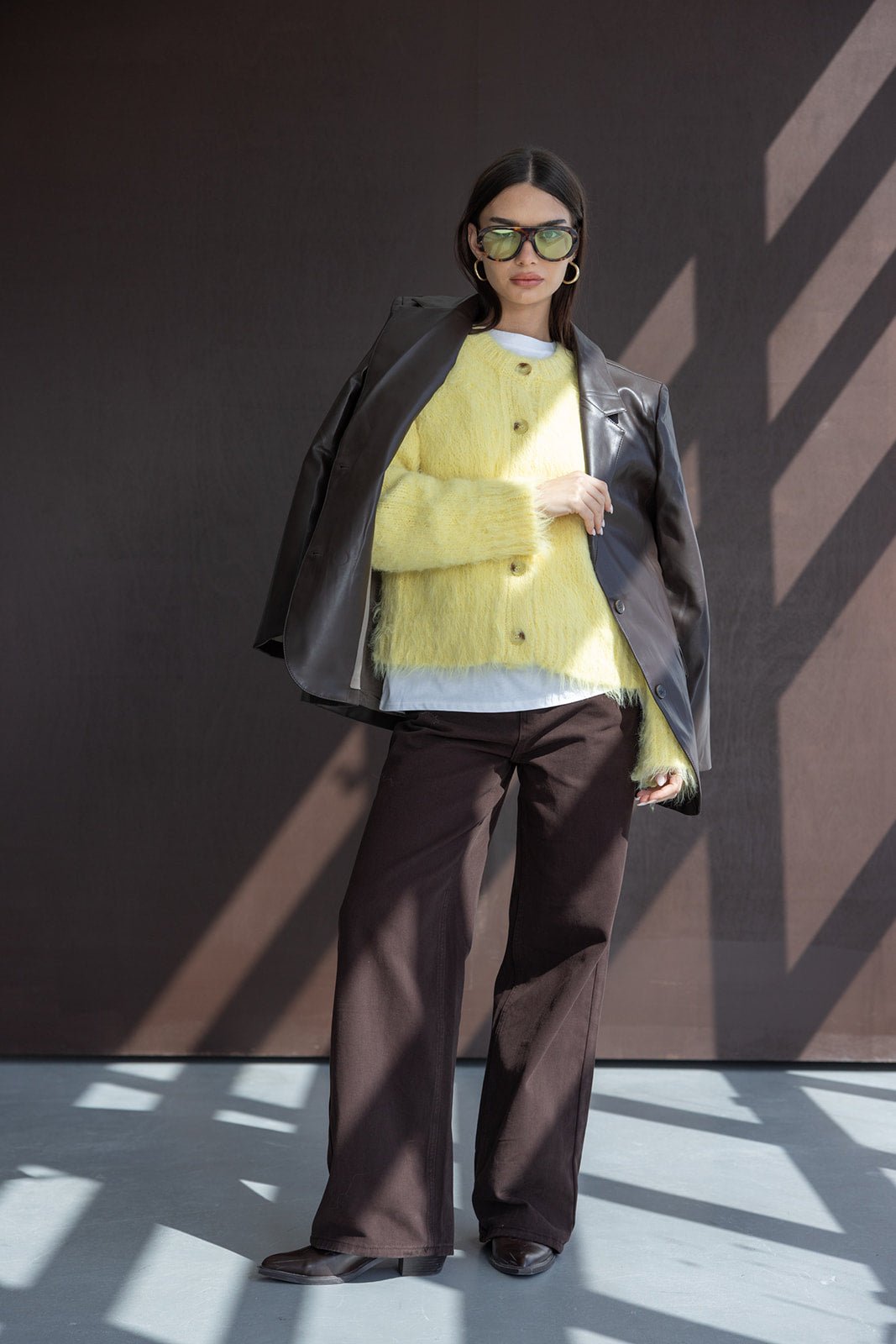 אישה לובשת מעיל עור שחור על כתפיה, סוודר צהוב פלפי, מכנסיים רחבים חומים ומשקפי שמש אופנתיים