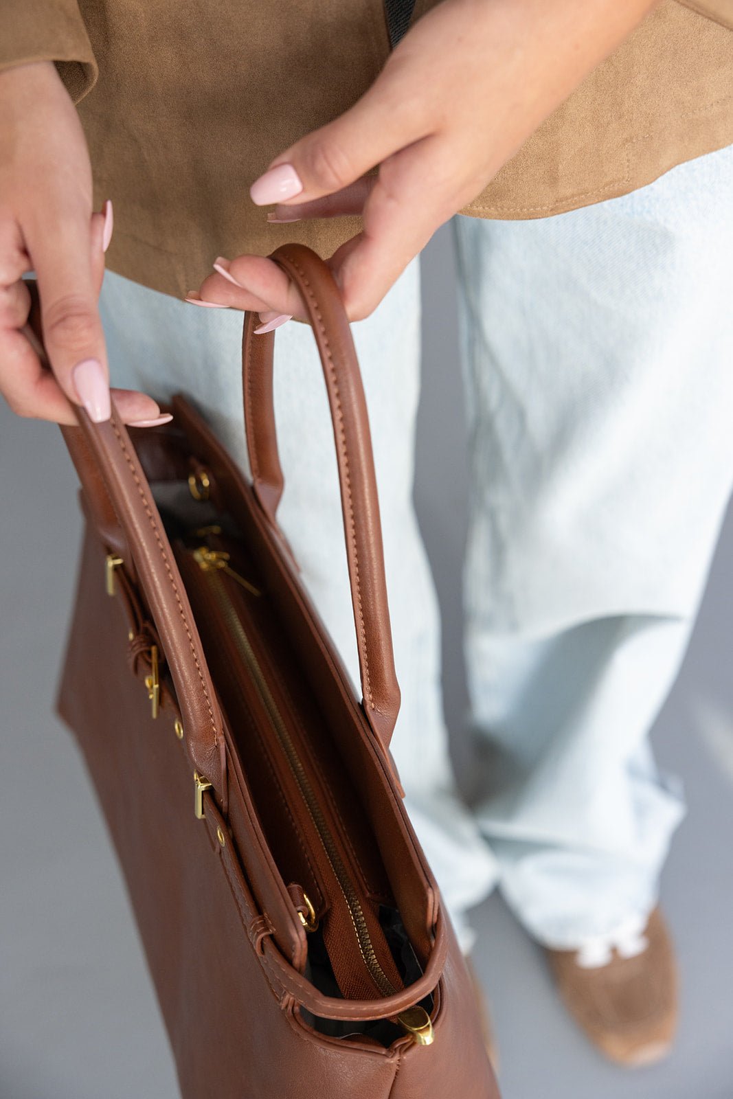 אישה מחזיקה תיק יד חום אלגנטי עם רוכסן, לבושה ג'ינס ונעלי סניקרס חומות אופנתיות